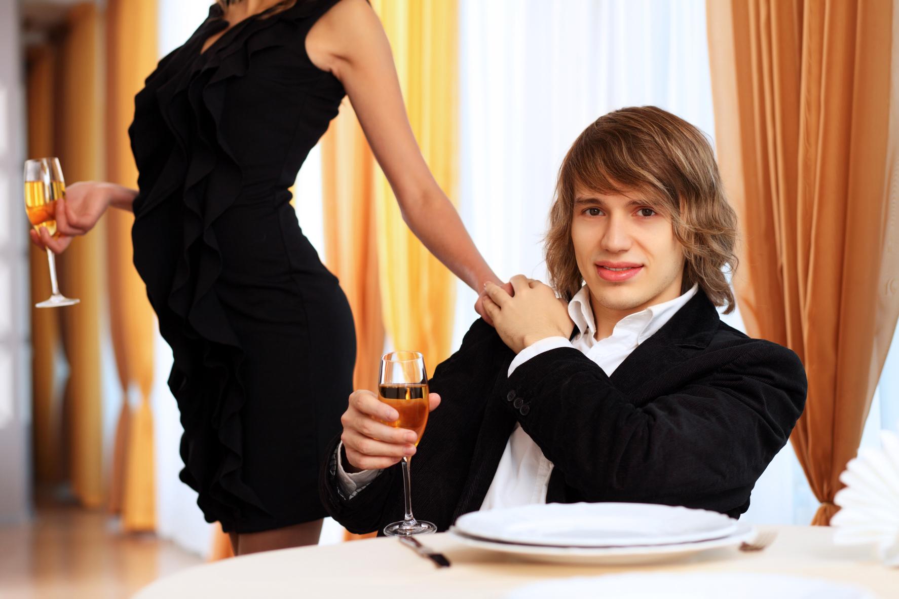 A white, long-haired man sitting at a table holding the hand of a woman who is standing in a tight, black dress