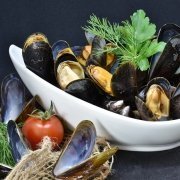 Tequila Mussels in a white bowl with a tomato on front and some green herbs behind the mussels in the dish