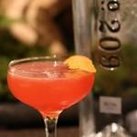 A Spring Gin Cocktail Recipe Made with Aperol and Love 2 orange cocktail in Champagne coupe with a lemon wedge garnish with a bottle of gin in the background