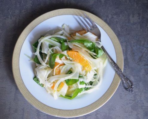 shaved fennel salad with citrus and arugula on a white plate with gold trim and a fork angled on the righthand side