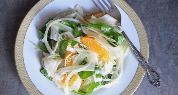 shaved fennel salad with citrus and arugula on a white plate with gold trim and a fork angled on the righthand side