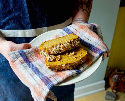 Make Sexy Desserts with Eat Cake Naked, an Aphrodisiac Cookbook 2 Pumpkin Coffee Cake from Eat Cake Naked being held by two hands on a white plate with a plaid tea towel