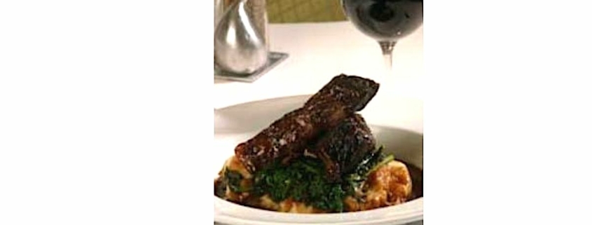 Closeup of red wine short ribs with figs in a white dish with a glass of wine and a silver fig in the backgorund