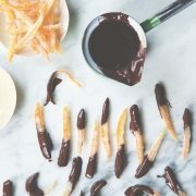 closeup of chocolate covered candied citrus peel on a pale blue background with a small pot of chocolate and extra orange peels on the side