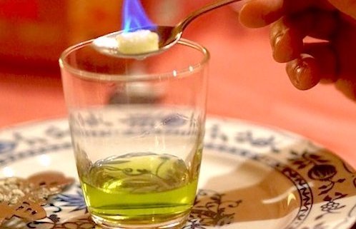 Absinthe Aphrodisiac Use in History 1 glass with absinthe and hand holding Absinthe spoon with a flaming sugar cube over the top to illustrate absinthe aphrodisiac