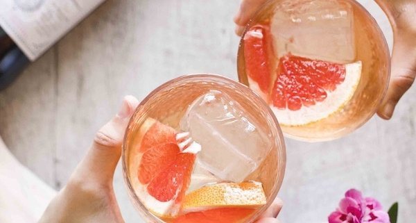 two hands, each holding a Lustau Spritz with a bottle of the vermouth in the background