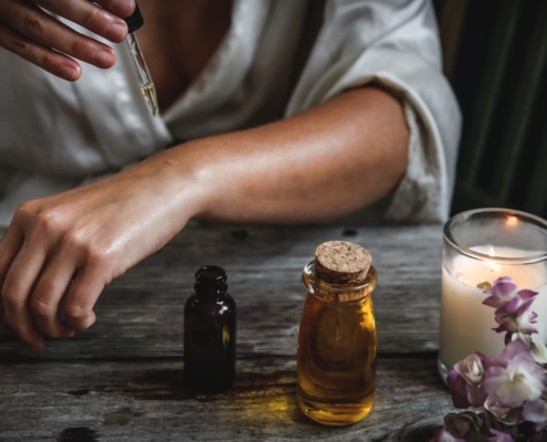 dark-skinned woman applying castor oil to her arm with a bottle of oil and a candle in the foreground to illustrate castor oil benefits to skin