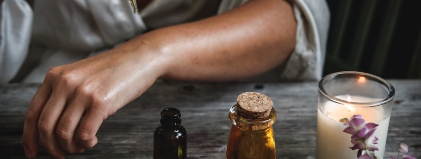 dark-skinned woman applying castor oil to her arm with a bottle of oil and a candle in the foreground to illustrate castor oil benefits to skin