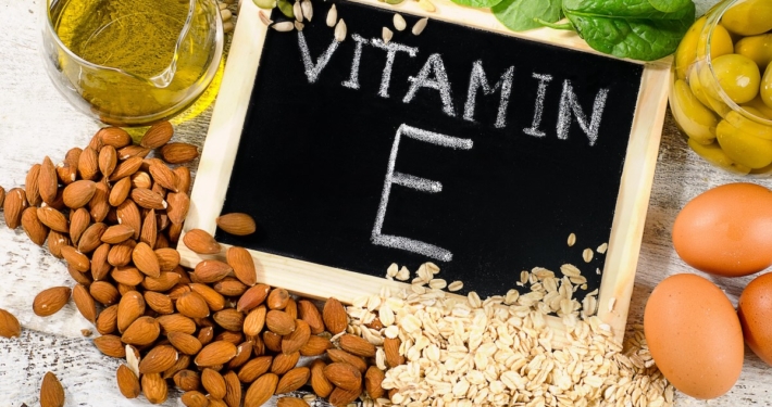 chalkboard with "vitamin E" written in white chalk, surrounded by ingredients rich in vitamin E including almonds, wheat germ oil