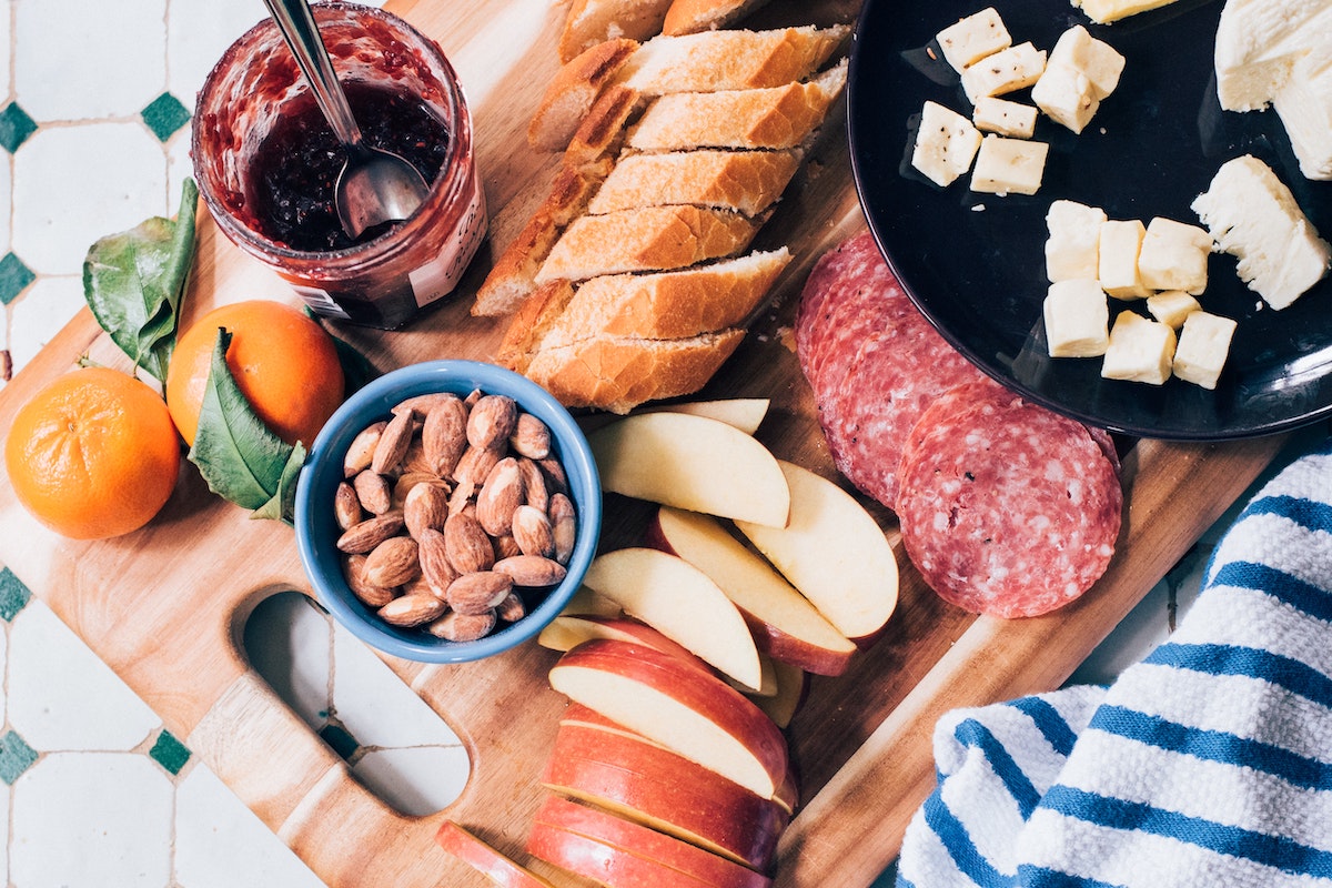 A traditional charcuterie board with sliced baguette, saussicon, apple slices, nuts and blueberry jam