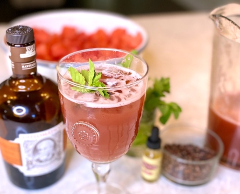 Watermelon cocktail next to a bottle of rum with a bowl of watermelon behind it and a pitcher of the rum punch on the side