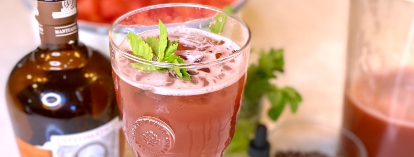 Watermelon cocktail next to a bottle of rum with a bowl of watermelon behind it and a pitcher of the rum punch on the side