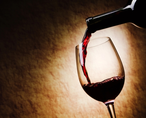 neck of a wine bottle with wine being poured into a stemmed glass on a spotlit, beige background