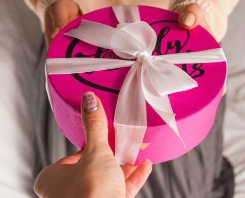 woman handing another person the gift of a round, pink cookie tin wrapped with a large, white bow