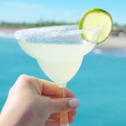Get Seriously Citrusy with the Best Patrón Citrónge Margarita Recipe 2 Woman's hand holding a margarita with a lime wheel garnish in front of a clear blue ocean