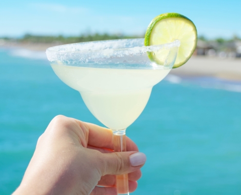 Woman's hand holding a margarita with a lime wheel garnish in front of a clear blue ocean