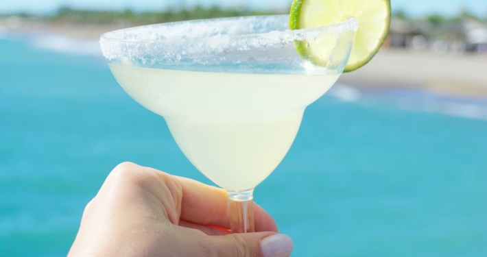 Woman's hand holding a margarita with a lime wheel garnish in front of a clear blue ocean
