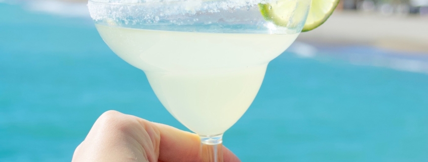 Woman's hand holding a margarita with a lime wheel garnish in front of a clear blue ocean
