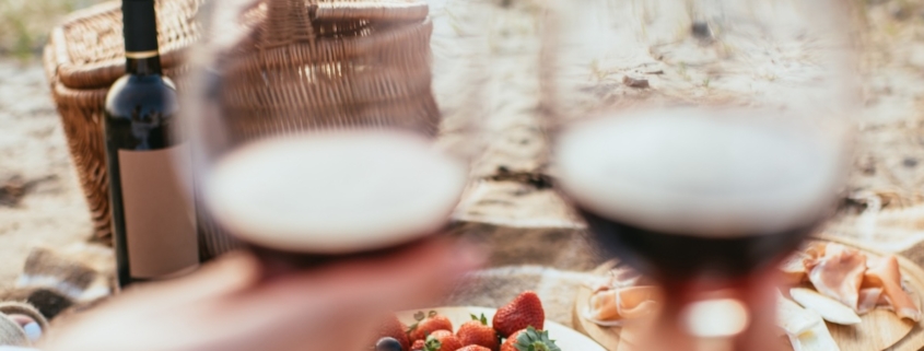 Couple's hands holding two blurred glasses of red wine in the foreground with the camera focused on a picnic blanket with assorted food in the background