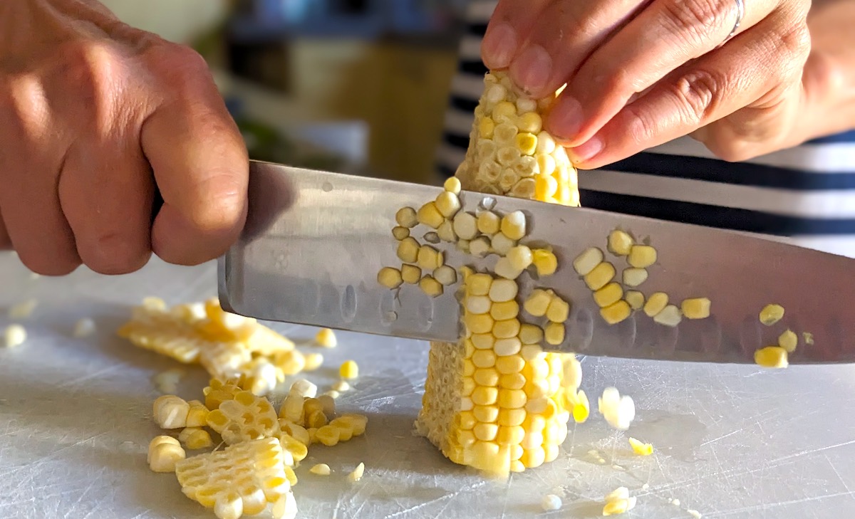 Hand holding a chef's knife illustrating a method to remove corn from the cob by cutting the cob in half first