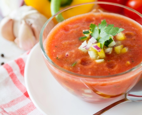 This Heirloom Tomato Gazpacho Is Summer in a Bowl 1 closeup of heirloom tomato gazpacho in a glass bowl on a red and white napkin with garlic, tomato and a yellow pepper in the background