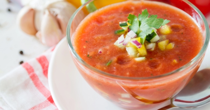 closeup of heirloom tomato gazpacho in a glass bowl on a red and white napkin with garlic, tomato and a yellow pepper in the background