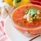 closeup of heirloom tomato gazpacho in a glass bowl on a red and white napkin with garlic, tomato and a yellow pepper in the background