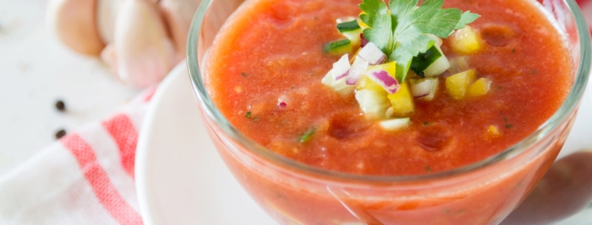 closeup of heirloom tomato gazpacho in a glass bowl on a red and white napkin with garlic, tomato and a yellow pepper in the background