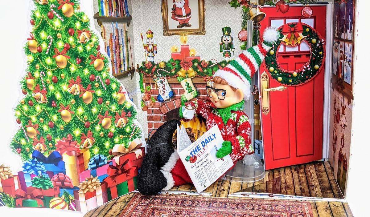 Sample World Elf Org scene with an Elf on the Shelf reading a newspaper next to a Christmas tree in a paper cutout house