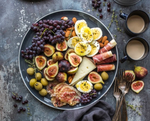 Bowl of organic ingredients including eggs, cheese, fruits and nuts on a dark background with two mugs and assorted cut fruit on the side