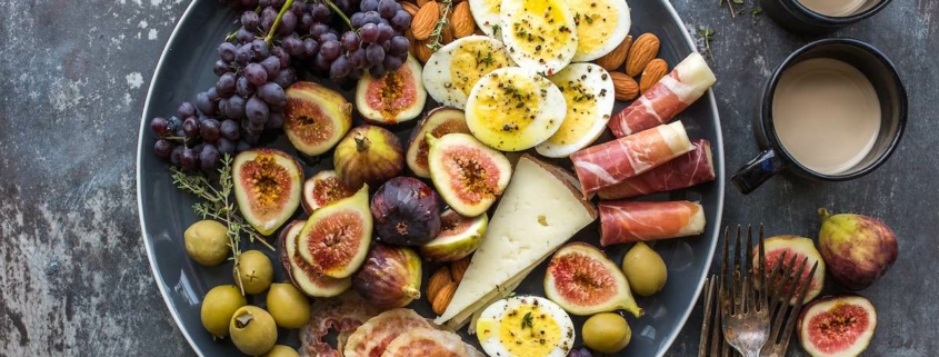 Bowl of organic ingredients including eggs, cheese, fruits and nuts on a dark background with two mugs and assorted cut fruit on the side