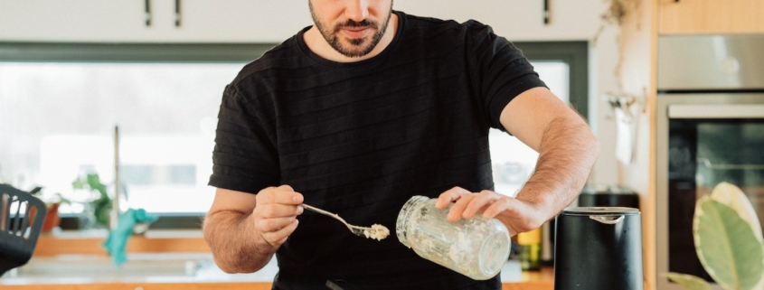 Man in a black shirt in a kitchen standing in front of a stove scooping something from a jar into a cast iron pan.