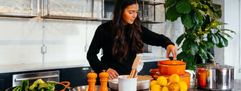 woman standing over a pot on the stove in a large, white kitchen with a bowl of lemons and a colander of kale in the foreground.