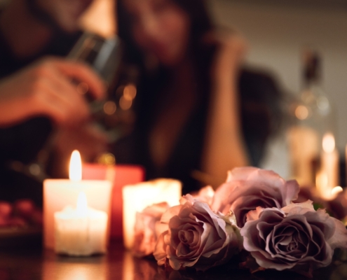 votive candles and a bouquet of roses in the foreground with the torsos of a couple snuggling and holding wine in the background