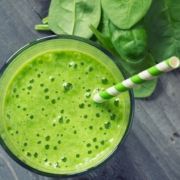 closeup of spinach and ginger smoothie with a green and white paper straw on a dark background with scattered spinach leaves