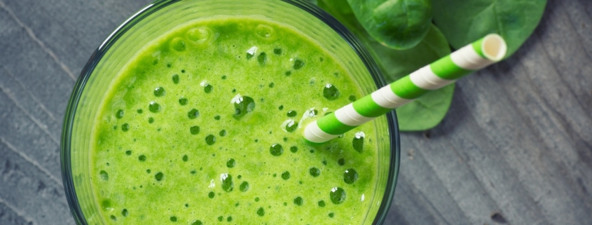 closeup of spinach and ginger smoothie with a green and white paper straw on a dark background with scattered spinach leaves