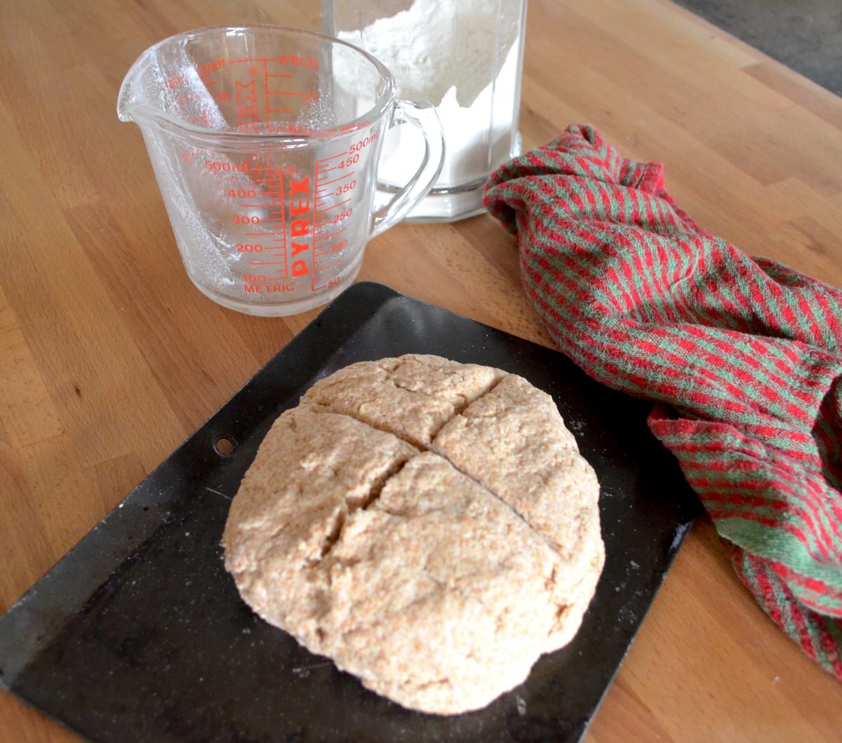 closeup of uncooked brown soda bread on a black baking tray with a tea towel and measuring cup in the background.
