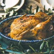 roast chicken in a blue roasting dish surrounded by olive branches on a table set for two with white candles burning in the background