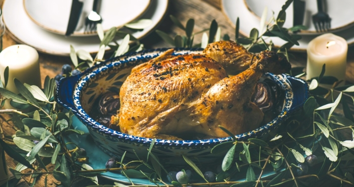 A Sexy Roast Chicken Recipe for Date Night 1 roast chicken in a blue roasting dish surrounded by olive branches on a table set for two with white candles burning in the background