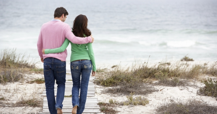 Man and woman walking arm-in-arm on a path through the dunes next to the ocean