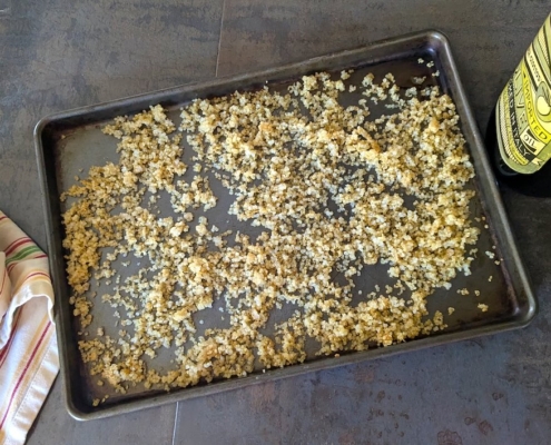 Crispy quinoa on a baking sheet with a white dish towel on the left and a bottle of olive oil and spoon on the right