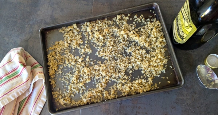 Crispy quinoa on a baking sheet with a white dish towel on the left and a bottle of olive oil and spoon on the right