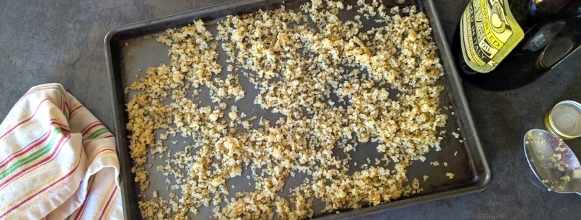 Crispy quinoa on a baking sheet with a white dish towel on the left and a bottle of olive oil and spoon on the right