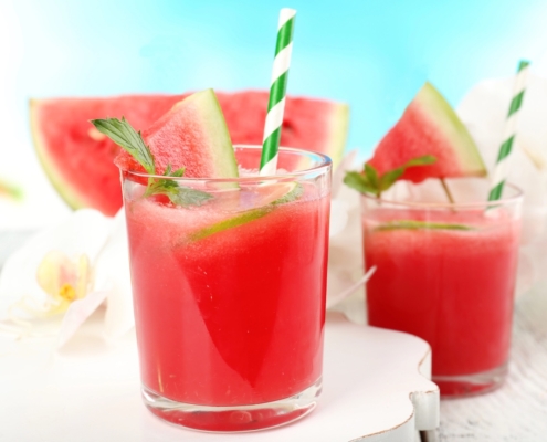 A Watermelon Cocktail With a Spicy Aphrodisac Twist 8 closeup of a watermelon cocktial with a blue and white striped paper straw and a sprig of mint with a second cocktail and watermelon slices in the background