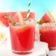 closeup of a watermelon cocktial with a blue and white striped paper straw and a sprig of mint with a second cocktail and watermelon slices in the background