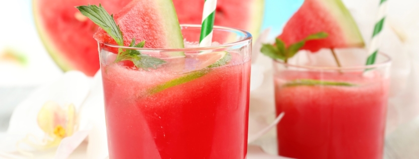 A Watermelon Cocktail With a Spicy Aphrodisac Twist 1 closeup of a watermelon cocktial with a blue and white striped paper straw and a sprig of mint with a second cocktail and watermelon slices in the background