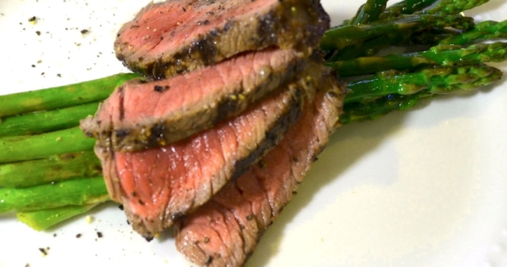 closeup of four slices of the marinated, grilled bison steak on top of asparagus on a white plate