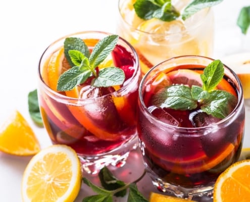 closeup of two glasses of red wine citrus sangria garnished with mint surrounded by cut lemons and oranges on a white background