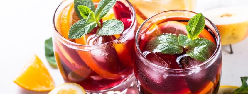 closeup of two glasses of red wine citrus sangria garnished with mint surrounded by cut lemons and oranges on a white background