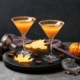 This Apple Pie Martini Tastes Like the Best Part of Apple Pie 4 two apple pie martinis on a black background surrounded by tiny pumpkins and cocktail making supplies